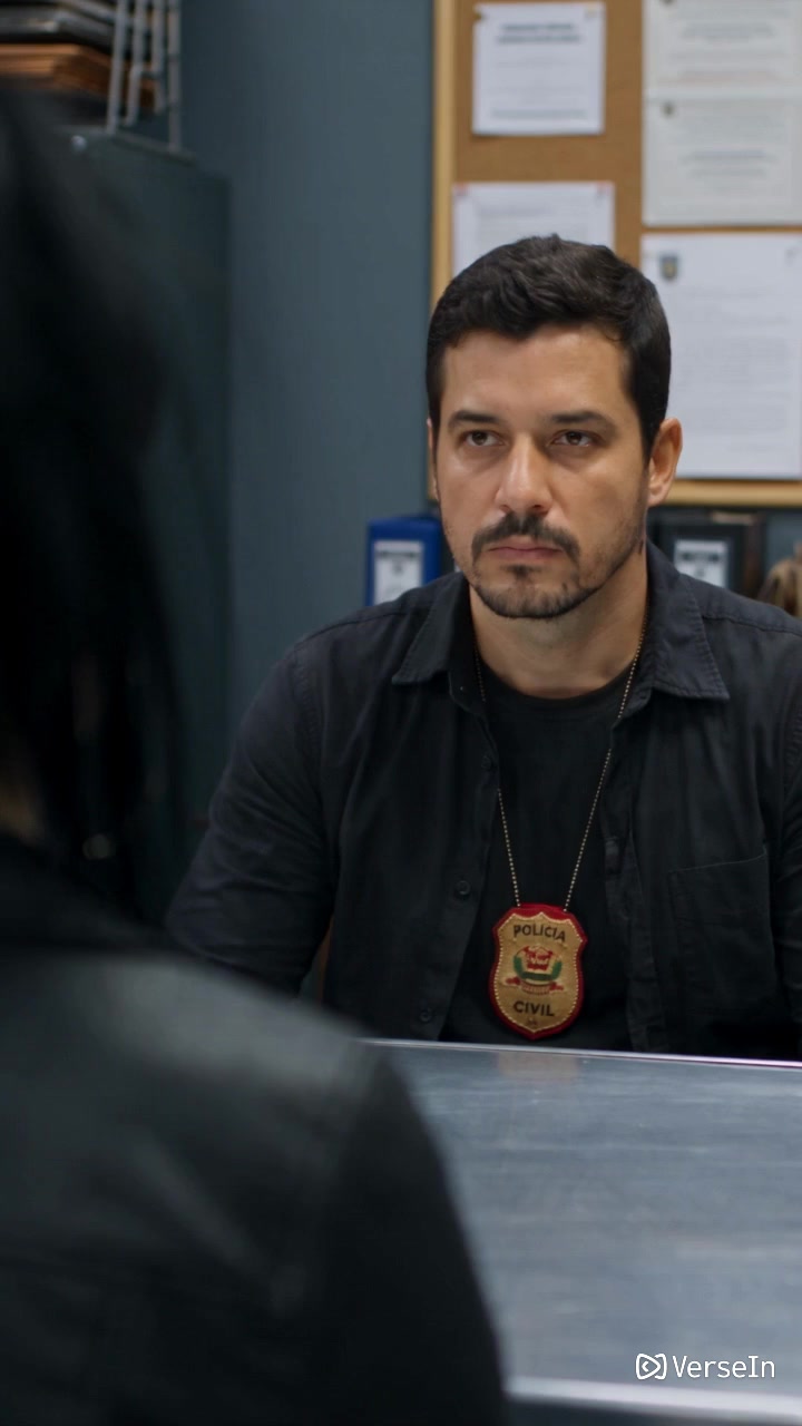 A detective wearing a badge sits across an interview table in a room, shown in a vertical frame.